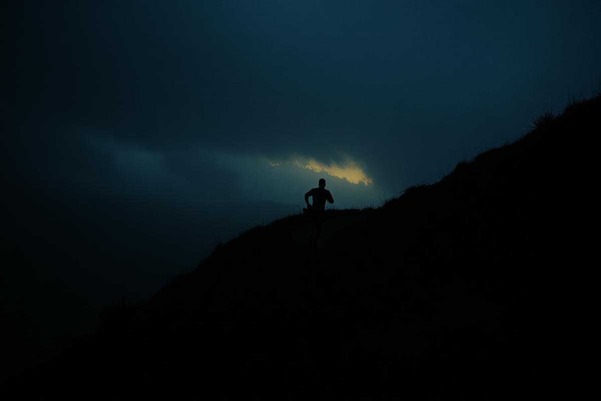 A lone runner climbing a steep dark hill under heavy storm clouds